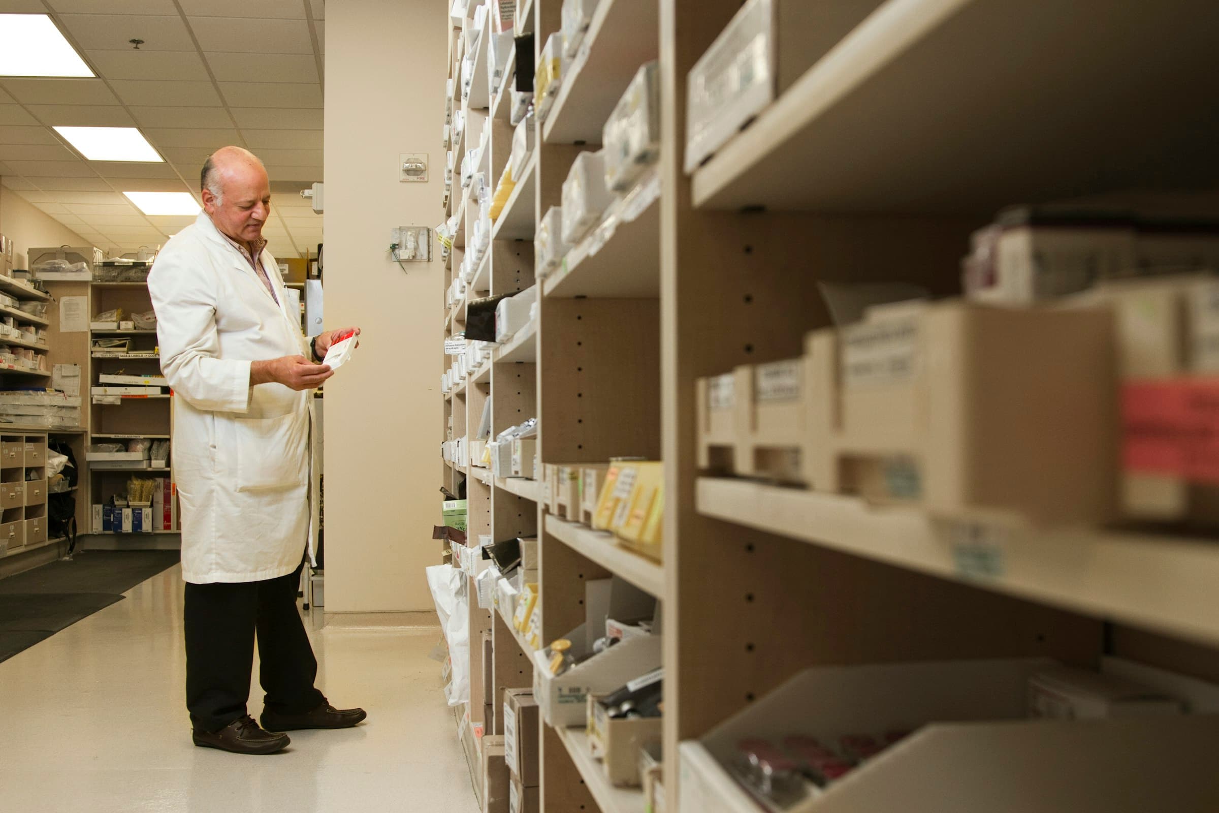 A pharmacist in a white coat reads a medication box in a hospital pharmacy aisle
