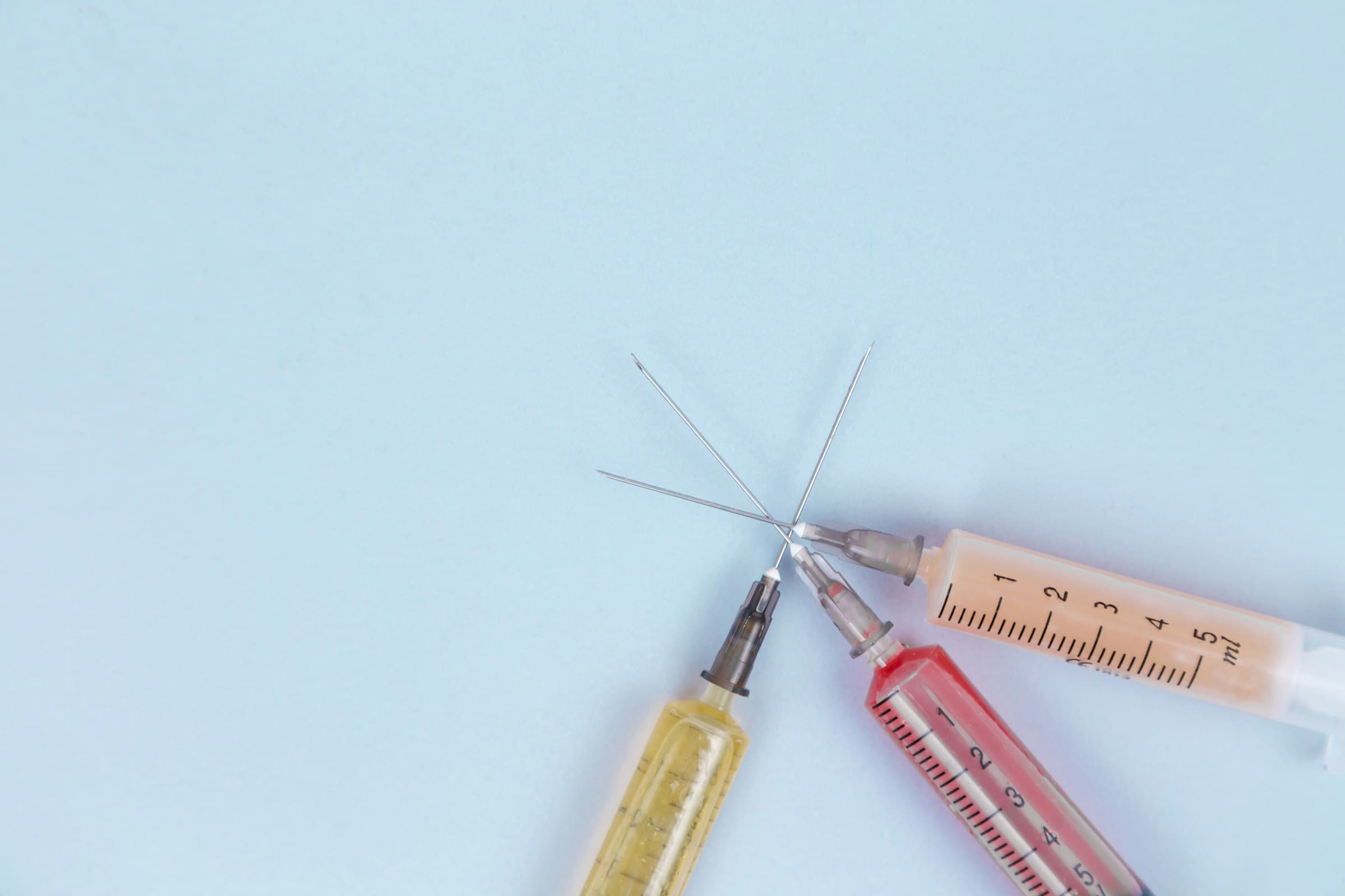 Three syringes with yellow, red and peach liquid on a pale blue surface